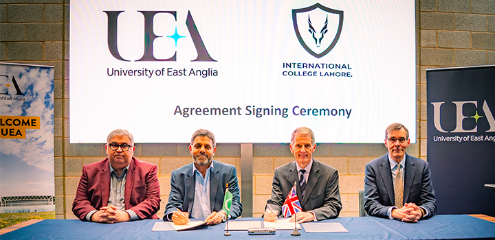 Four people signing documents seated at a blue table, two small flags in the middle of them and a large white screen is behind them.
