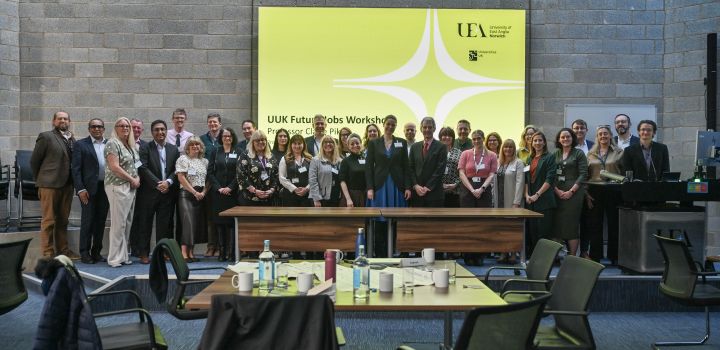 A large group of around 30 people stand behind a desk and in front of a large screen which bears the UEA logo and says UUK Future Jobs Workshop