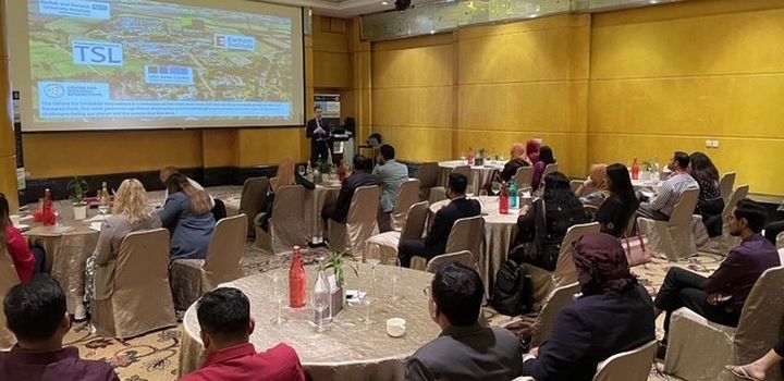 Groups of people at round tables in a conference room watch a projector screen with information about Norwich Research Park