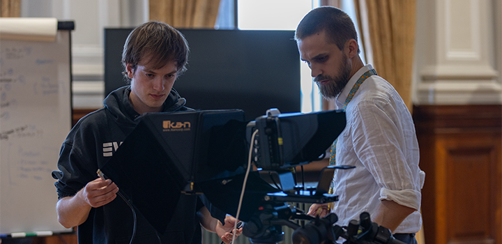 Two people are looking down at a news camera, one wearing a black hoodie and the other in a white shirt with a lanyard
