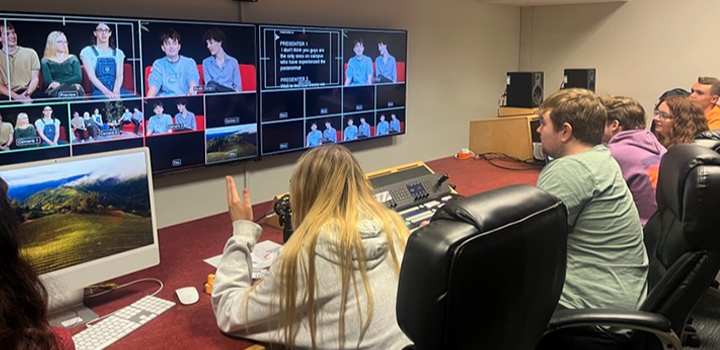 People in a control room monitor multiple screens showing a live broadcast. A woman gestures while others focus on the displays.