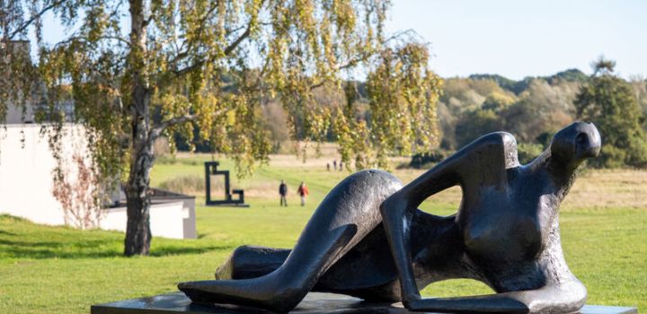 Modern sculpture by Henry Moore outside the Sainsbury Centre of a reclining figure on a lawn with trees and people walking in the background on a sunny day.