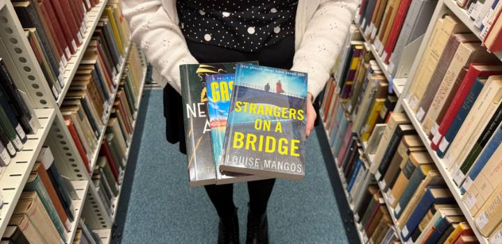 A woman holding three books in a library aisle, with 'Strangers on a Bridge' by Louise Mangos visible. Shelves filled with books surround them.