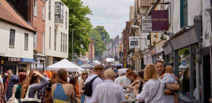 The Norwich Lanes street fair, with people walking, chatting, and shopping. Stalls line the street, surrounded by historic buildings and greenery.