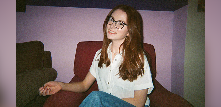 Daisy Innes smiling, with long hair and glasses as she sits on a red chair. She is wearing a white shirt and jeans, against a purple wall