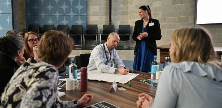 A group of people are having a debate around a table. One man is writing notes of what is said on a large piece of paper