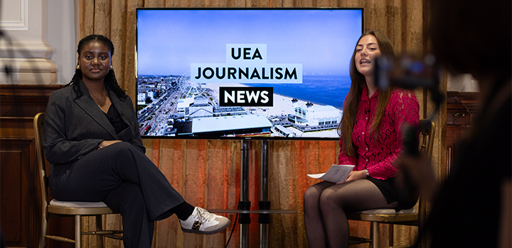 Two people sitting on chairs, one wearing a black suit and the other wearing a red top and skirt; a screen displaying 'UEA JOURNALISM NEWS' is in the middle of them