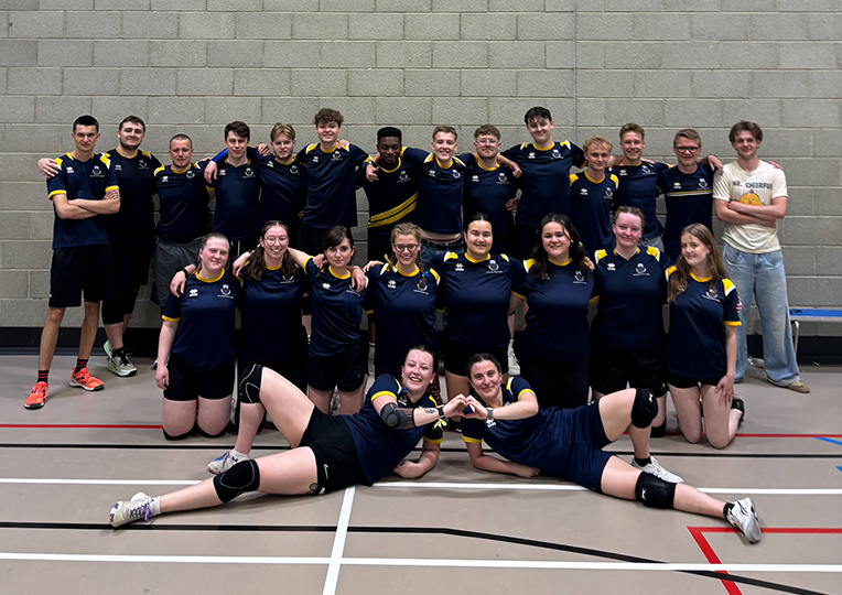 A group of people in matching sports uniforms pose together in a gym, with two people lying in front making a heart shape with their hands