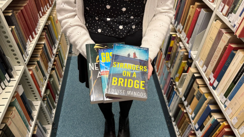 A person standing in a library aisle holding three books toward the camera. The front book is "Strangers on a Bridge" by Louise Mangos.