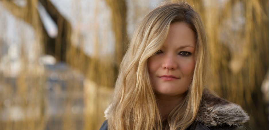 A blonde haired woman is looking at the camera, wearing a fur-collared jacket outdoors.