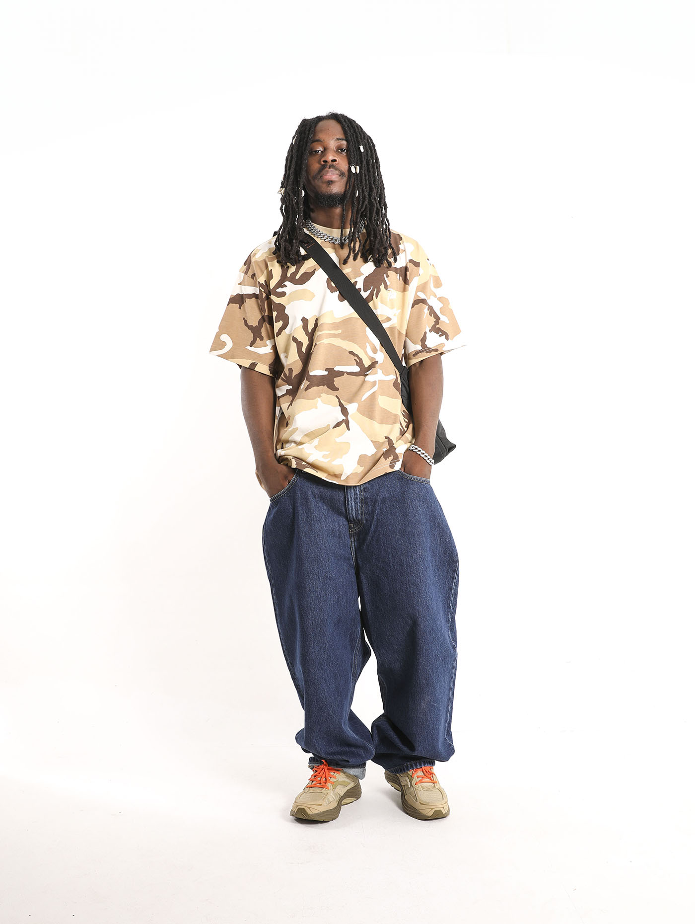 Person with dreadlocks wearing a camouflage shirt, baggy jeans, and sneakers, standing against a plain white background.