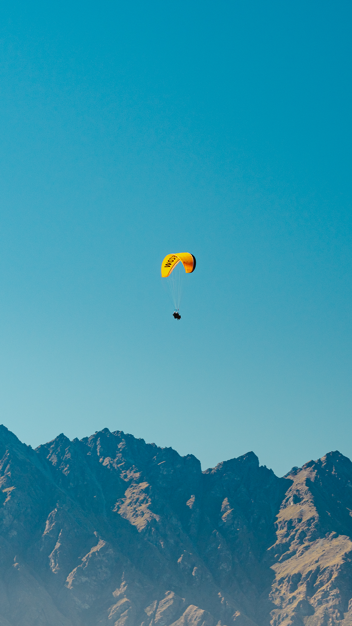 A yellow paraglider soars above rugged, sunlit mountains under a clear blue sky.