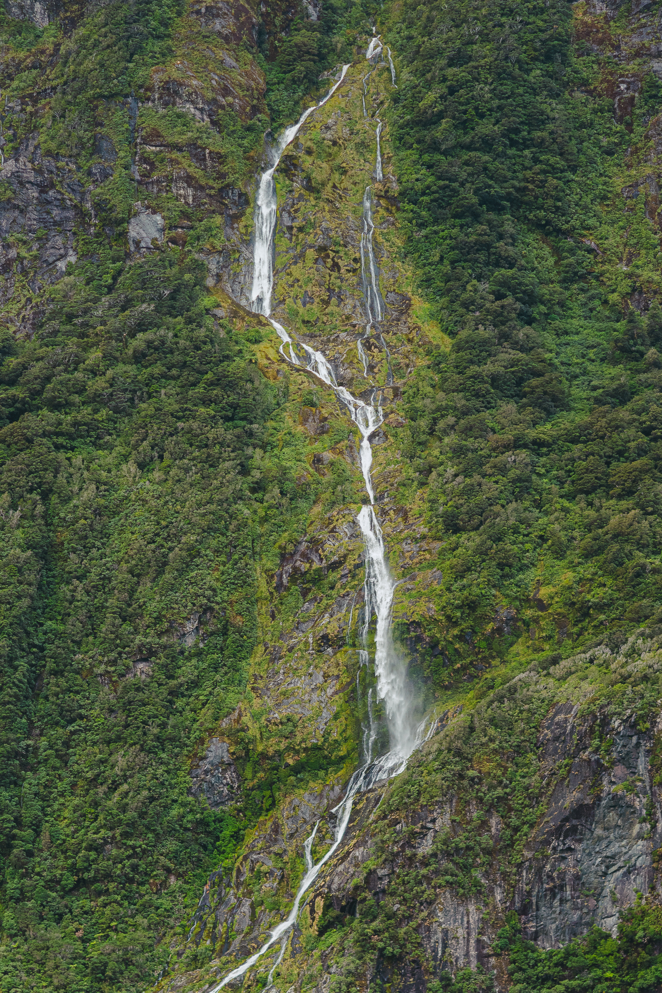 A tall, narrow waterfall cascades down a lush, green mountainside into a turquoise lake below.