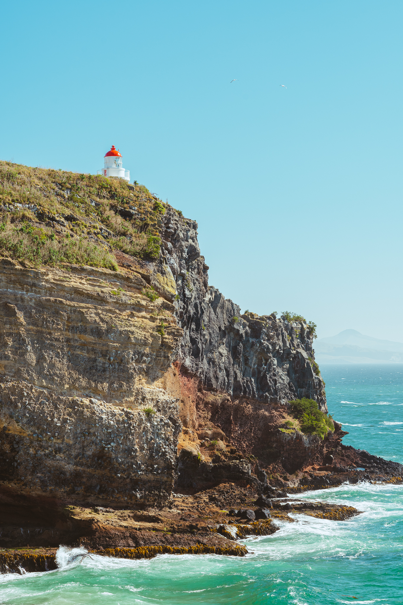 A small lighthouse with a red roof sits atop a steep, rocky cliff overlooking the ocean under a clear blue sky.