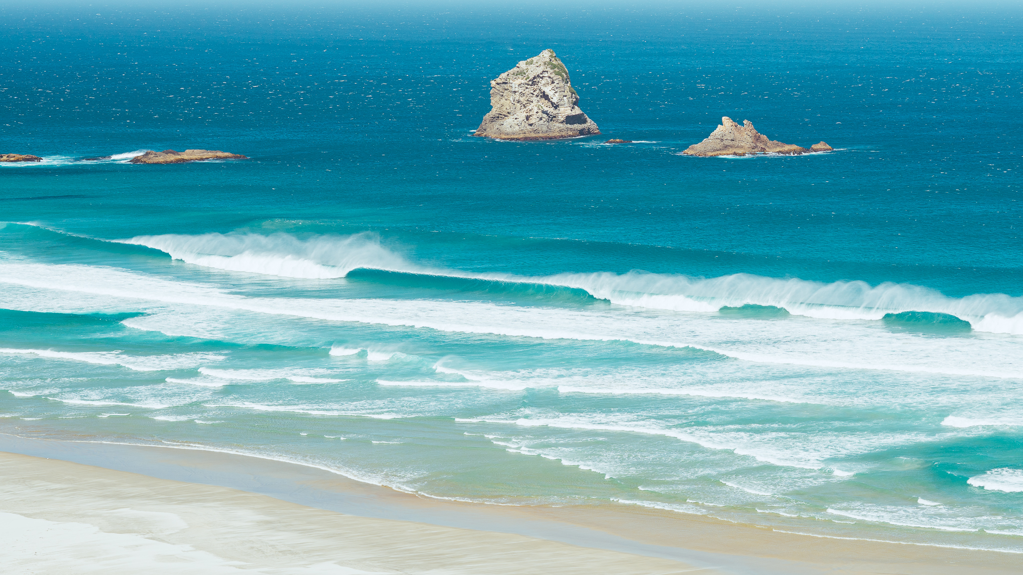 Two rocky islets in a vibrant blue ocean, with waves gently crashing onto a sandy beach under a clear sky.