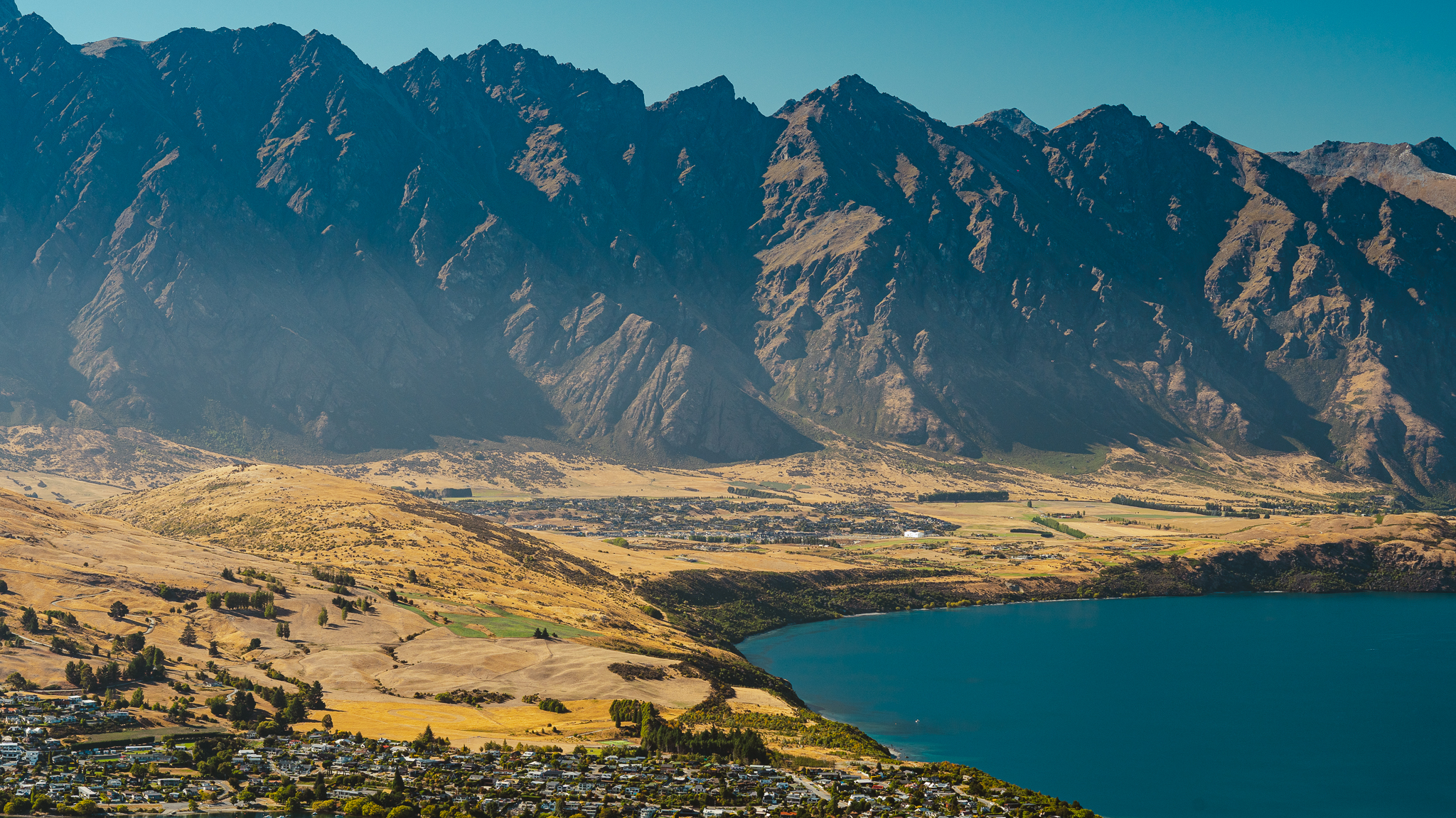 Aerial view of a lake with a mountainous backdrop, clear blue water, and a small town nestled along the shoreline under a bright blue sky.