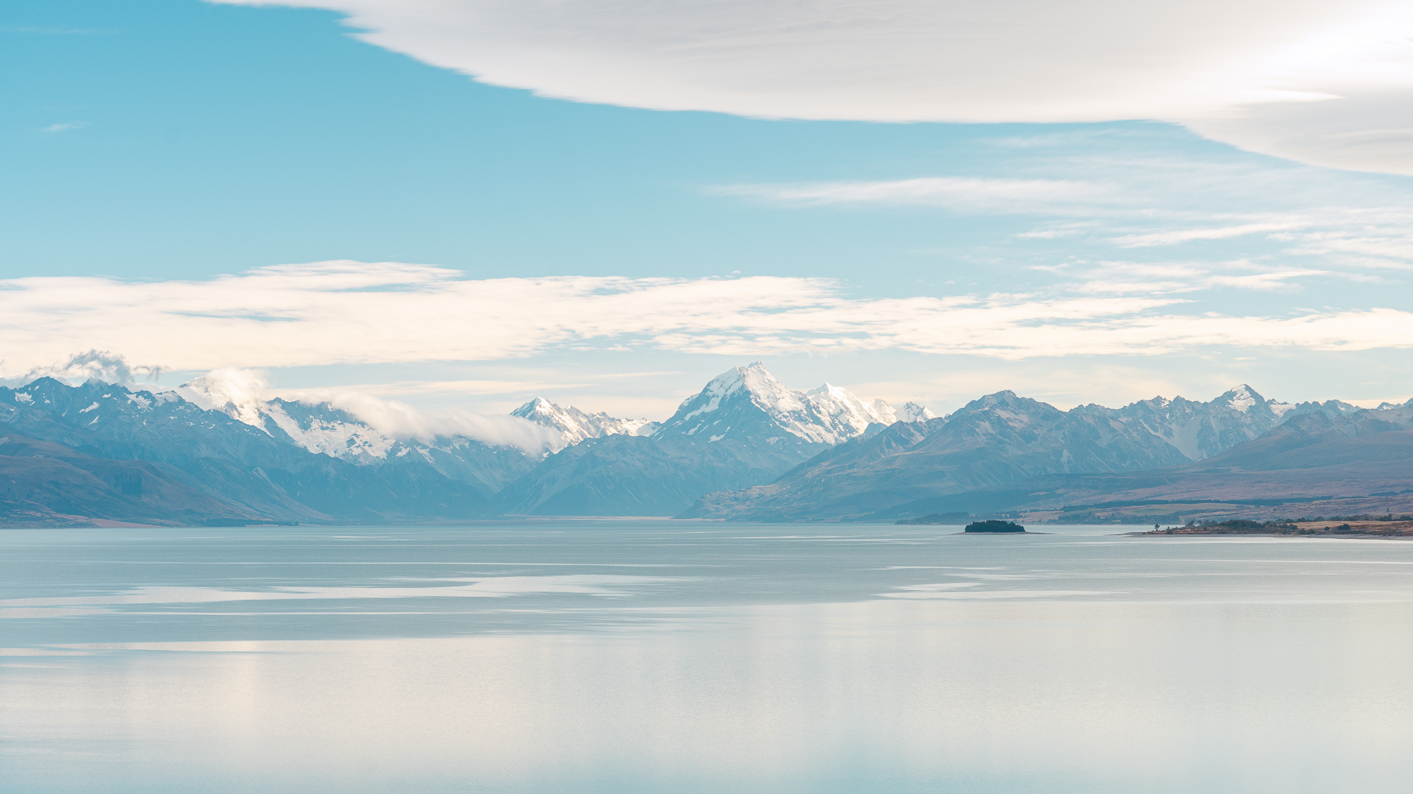 Serene landscape of snow-capped mountains and a calm lake under a blue sky with scattered clouds.