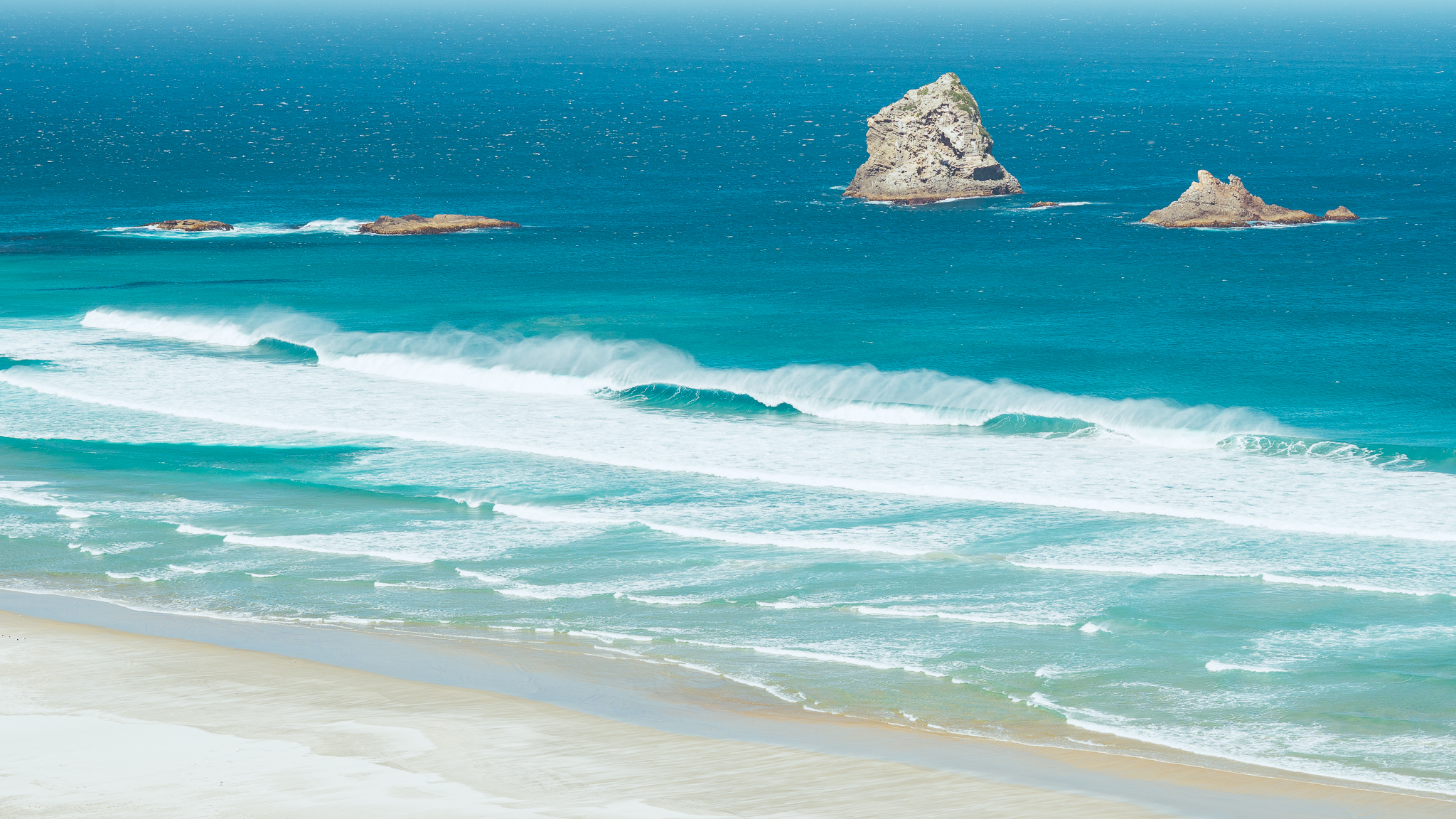 Two rocky islets in a vibrant blue ocean, with waves gently crashing onto a sandy beach under a clear sky.