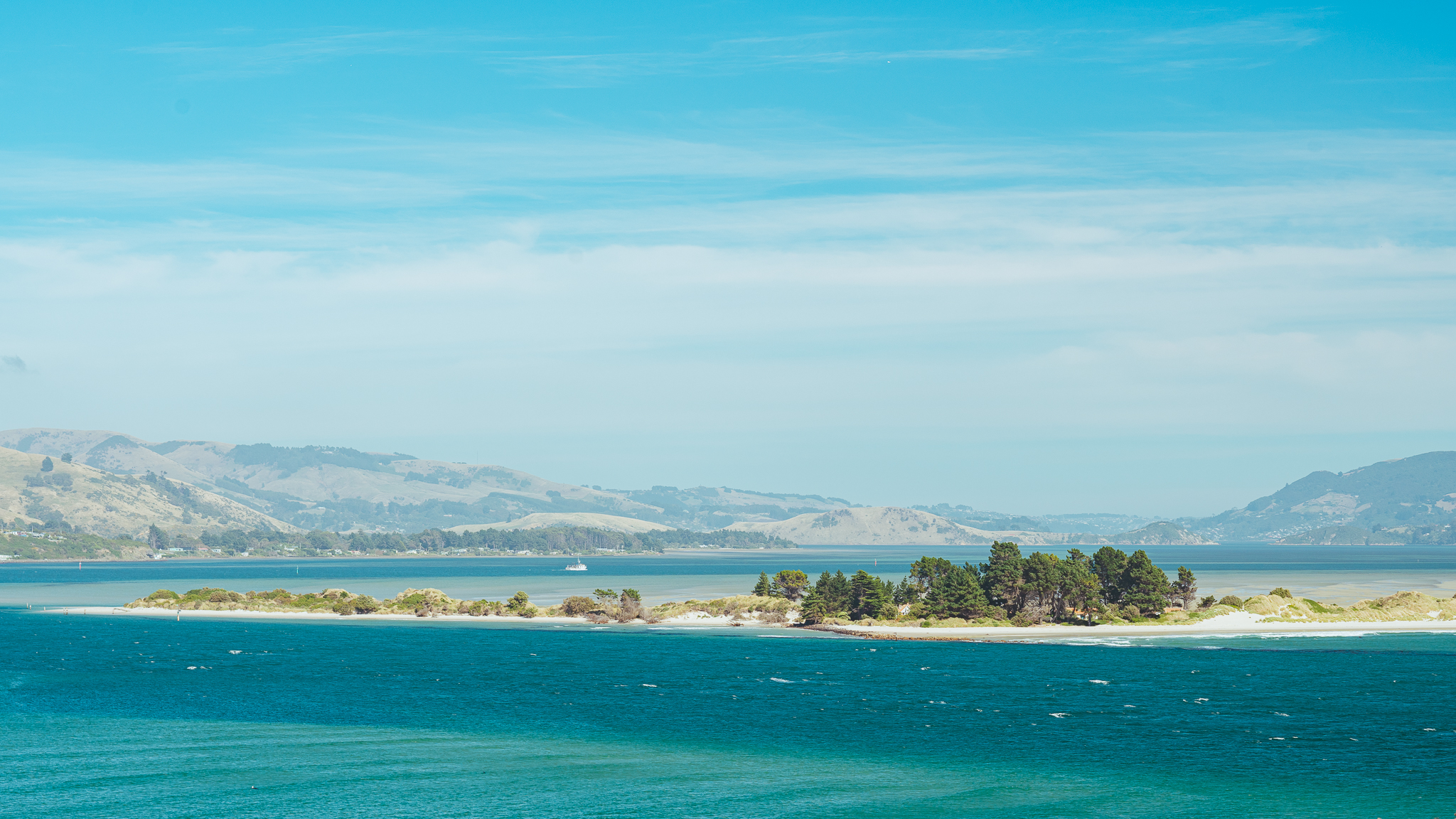 A small island with trees surrounded by turquoise water under a blue sky, with distant hills in the background.