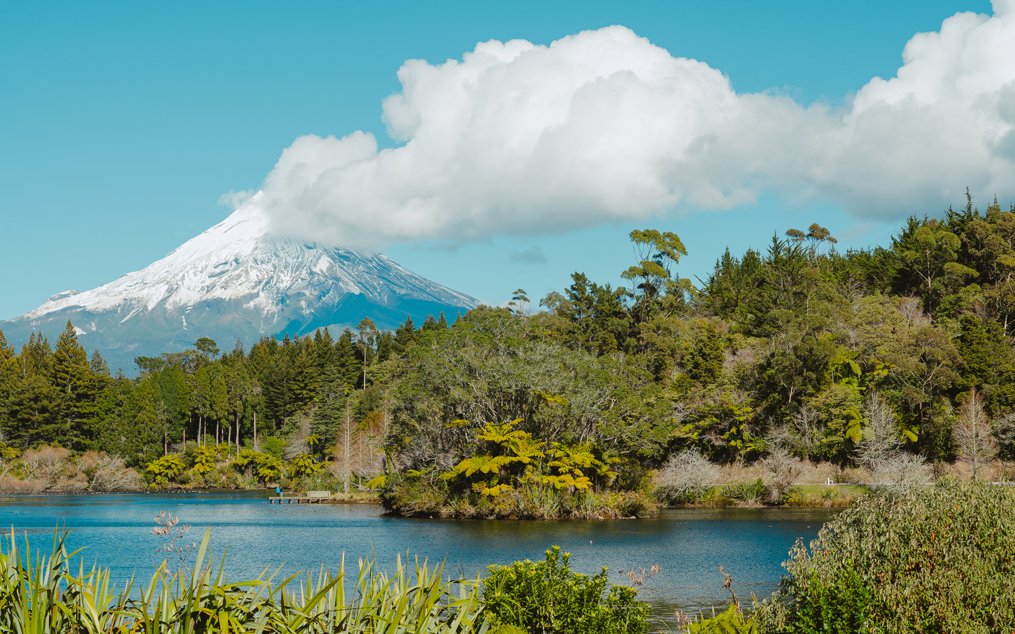 Snow-capped Mou with a cloud at the peak, surrounded by lush green forest and a tranquil lake in the foreground under a clear blue sky.