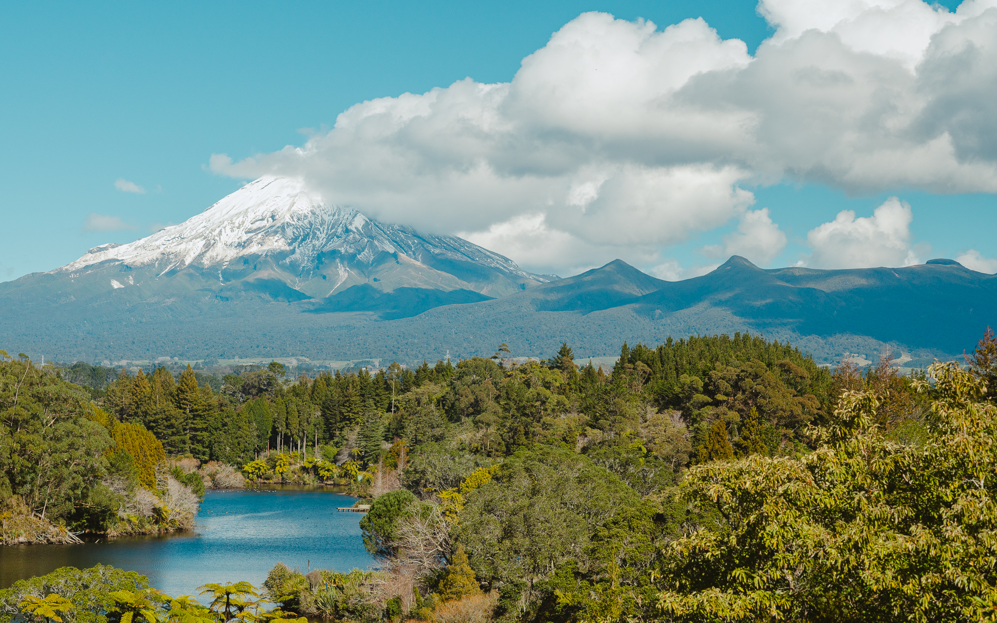 Snow-capped Mou with a cloud at the peak, surrounded by lush green forest and a tranquil lake in the foreground under a clear blue sky.