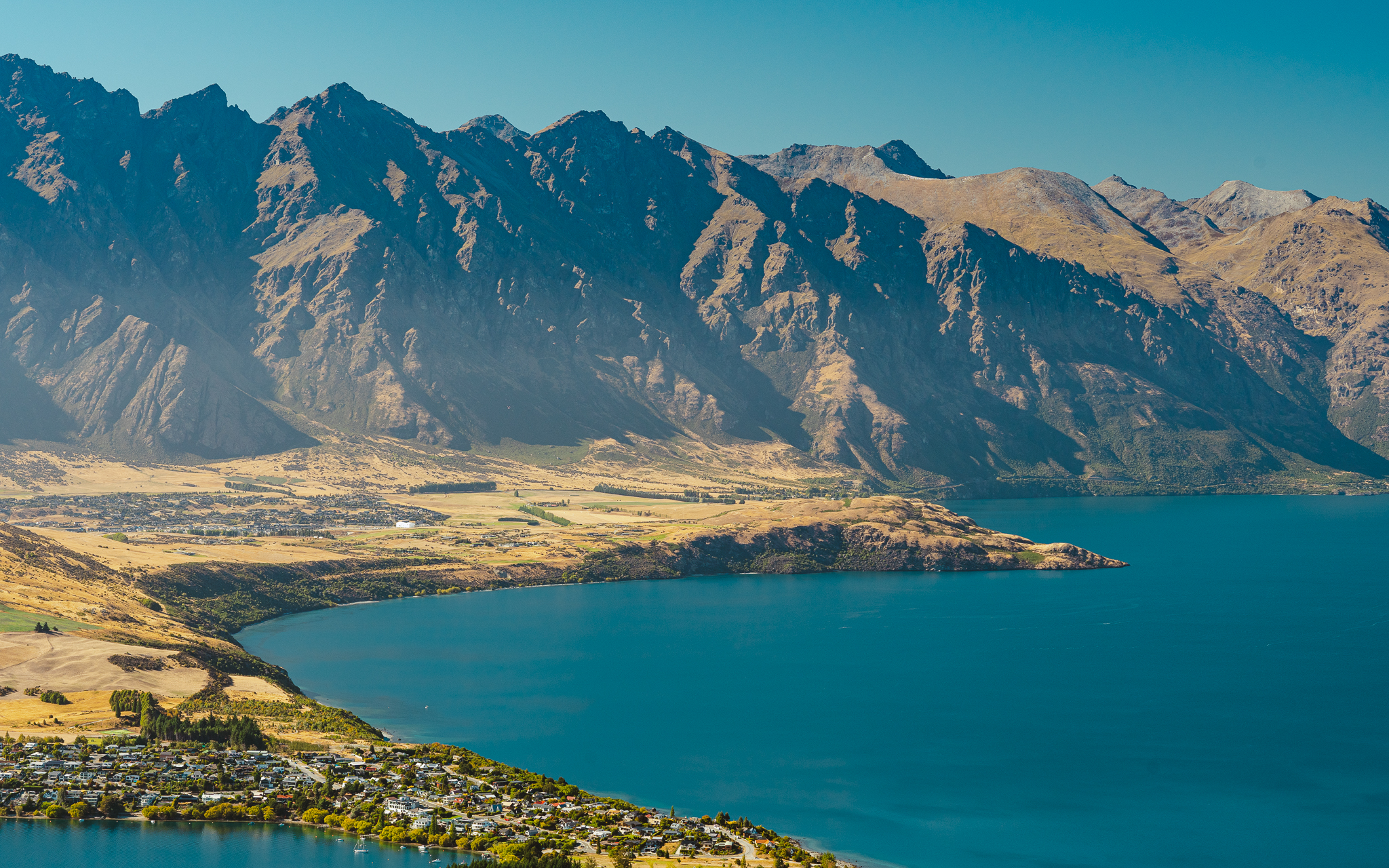 Aerial view of a lake with a mountainous backdrop, clear blue water, and a small town nestled along the shoreline under a bright blue sky.
