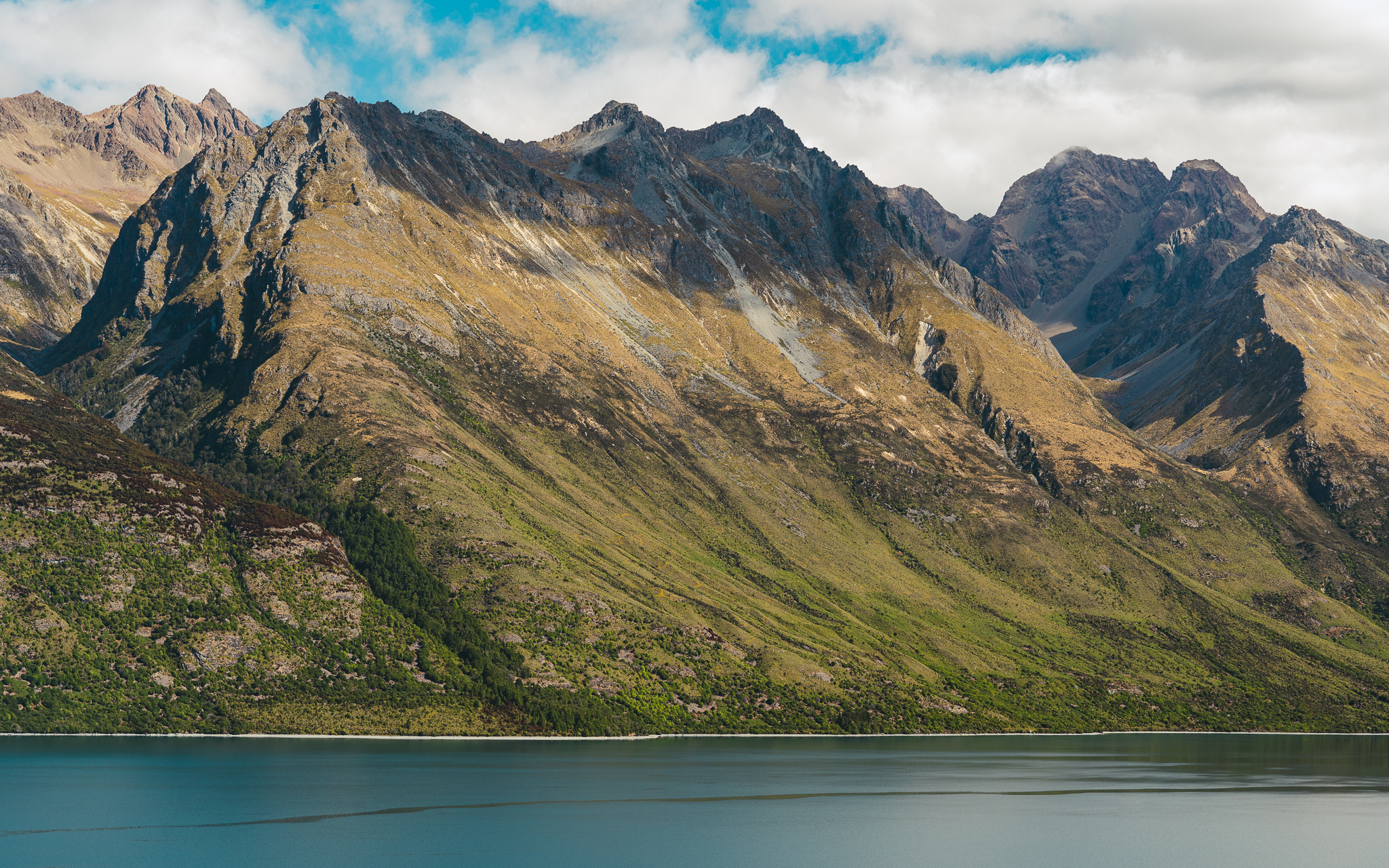 Majestic mountain range with rocky peaks and green slopes, bordering a calm blue lake under a partly cloudy sky.