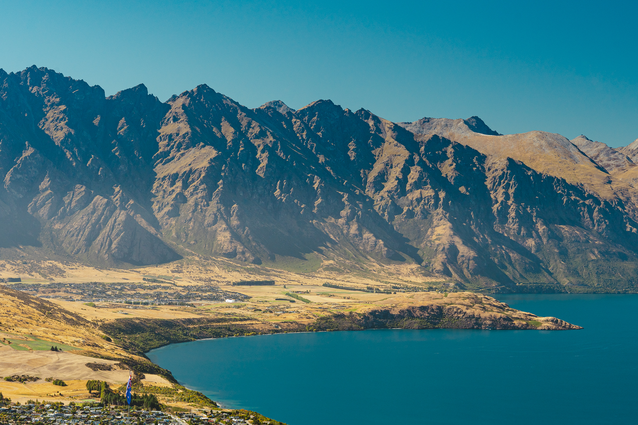 Aerial view of a lake with a mountainous backdrop, clear blue water, and a small town nestled along the shoreline under a bright blue sky.