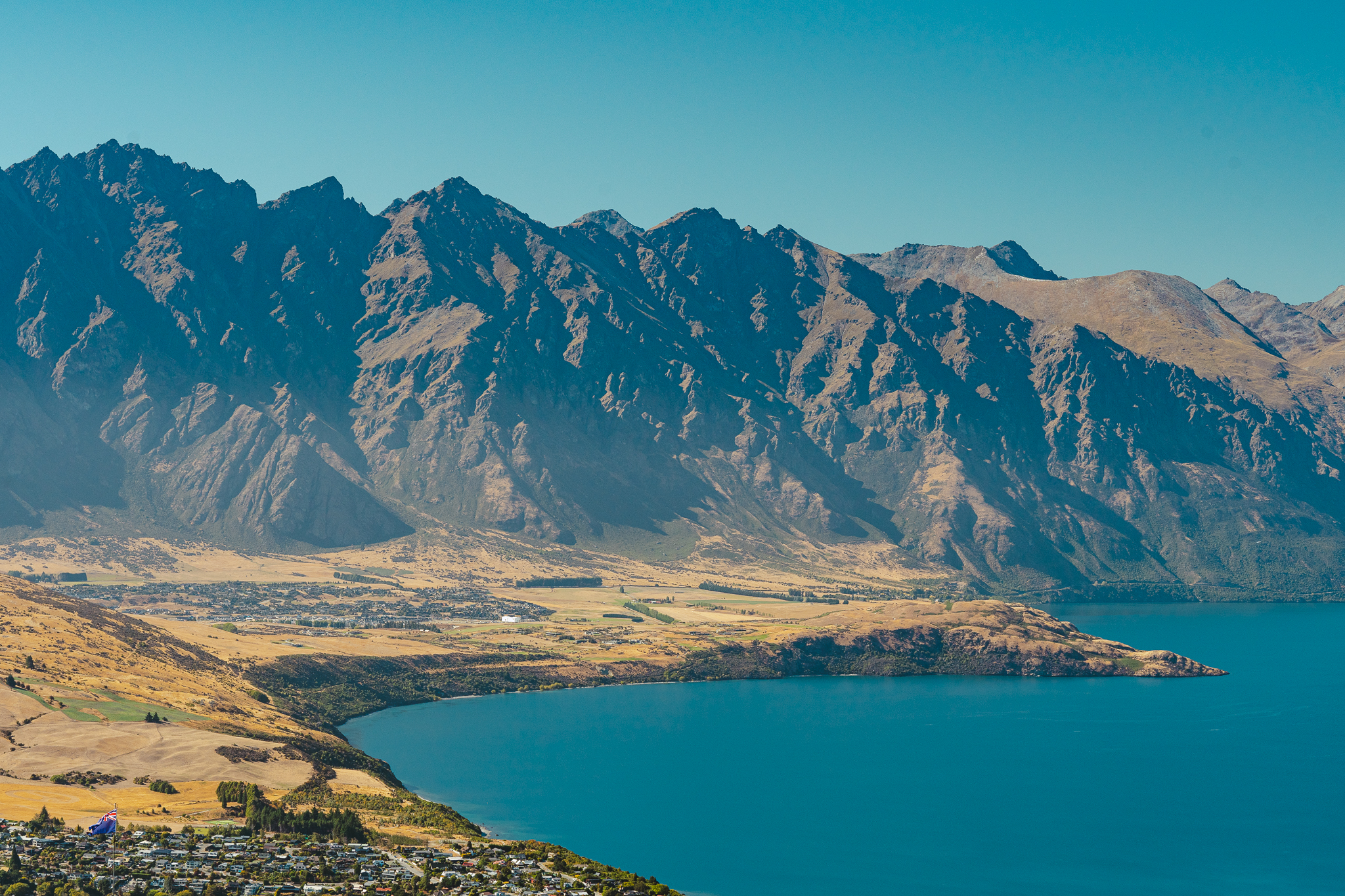 Aerial view of a lake with a mountainous backdrop, clear blue water, and a small town nestled along the shoreline under a bright blue sky.