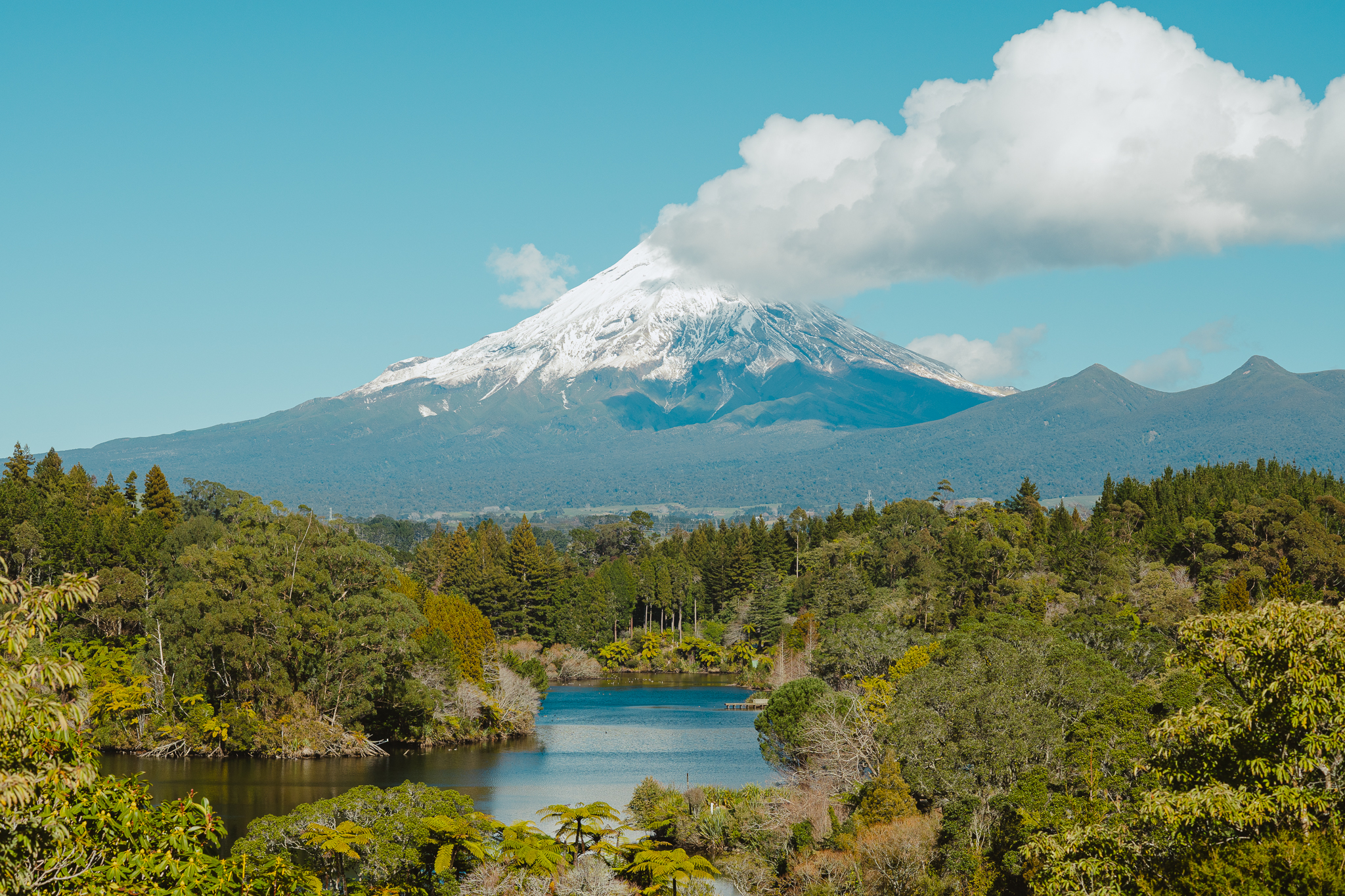 Snow-capped Mou with a cloud at the peak, surrounded by lush green forest and a tranquil lake in the foreground under a clear blue sky.