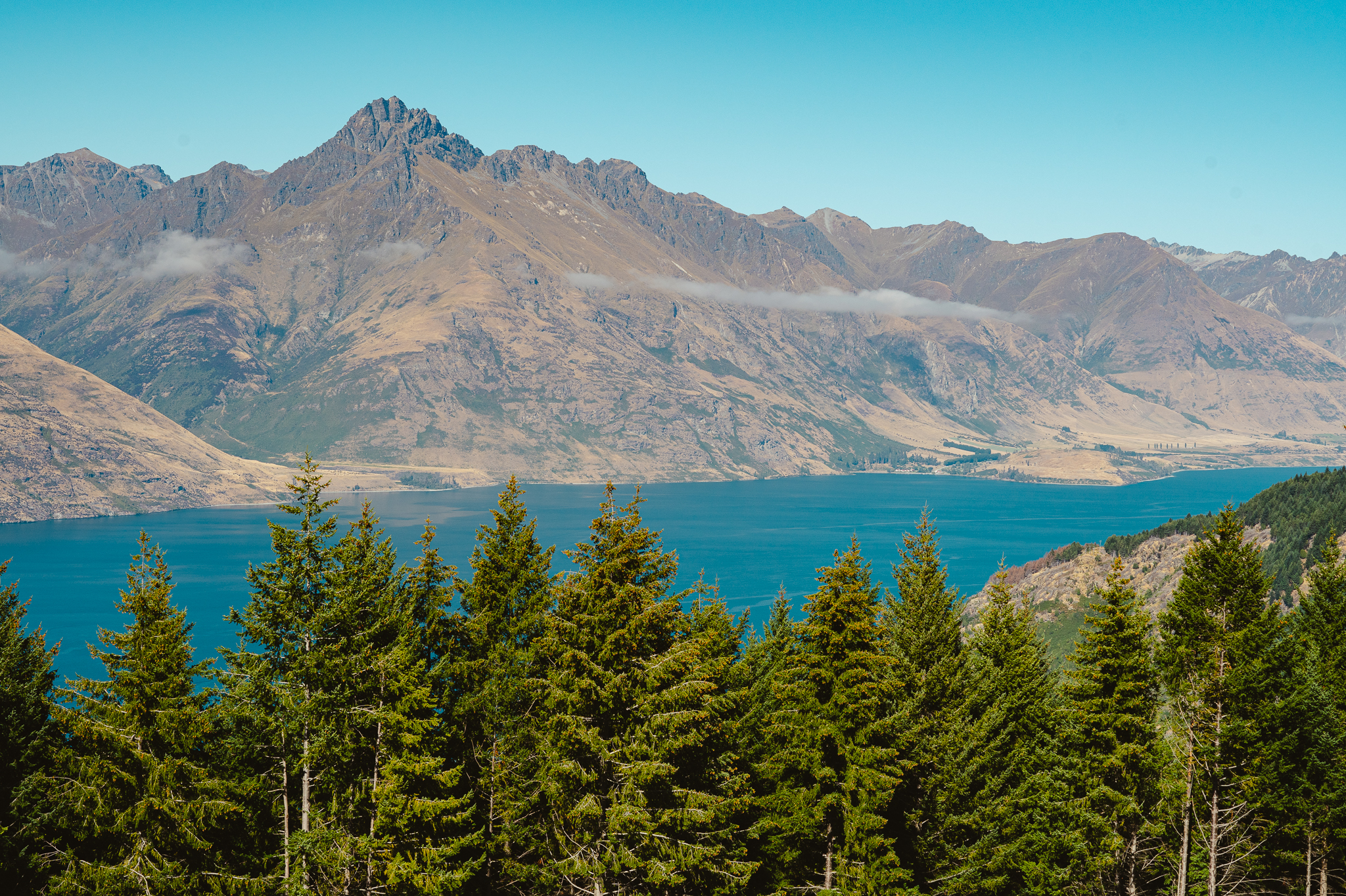 Mountain range and clear blue lake viewed from a forested area with evergreen trees under a clear blue sky.