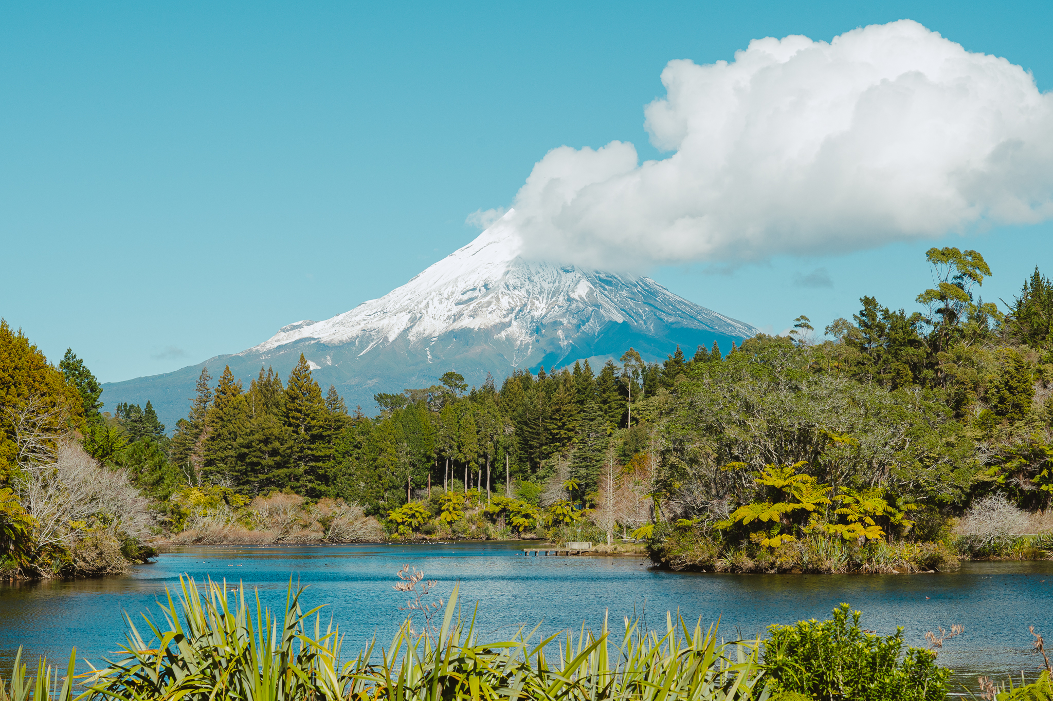 Snow-capped Mou with a cloud at the peak, surrounded by lush green forest and a tranquil lake in the foreground under a clear blue sky.