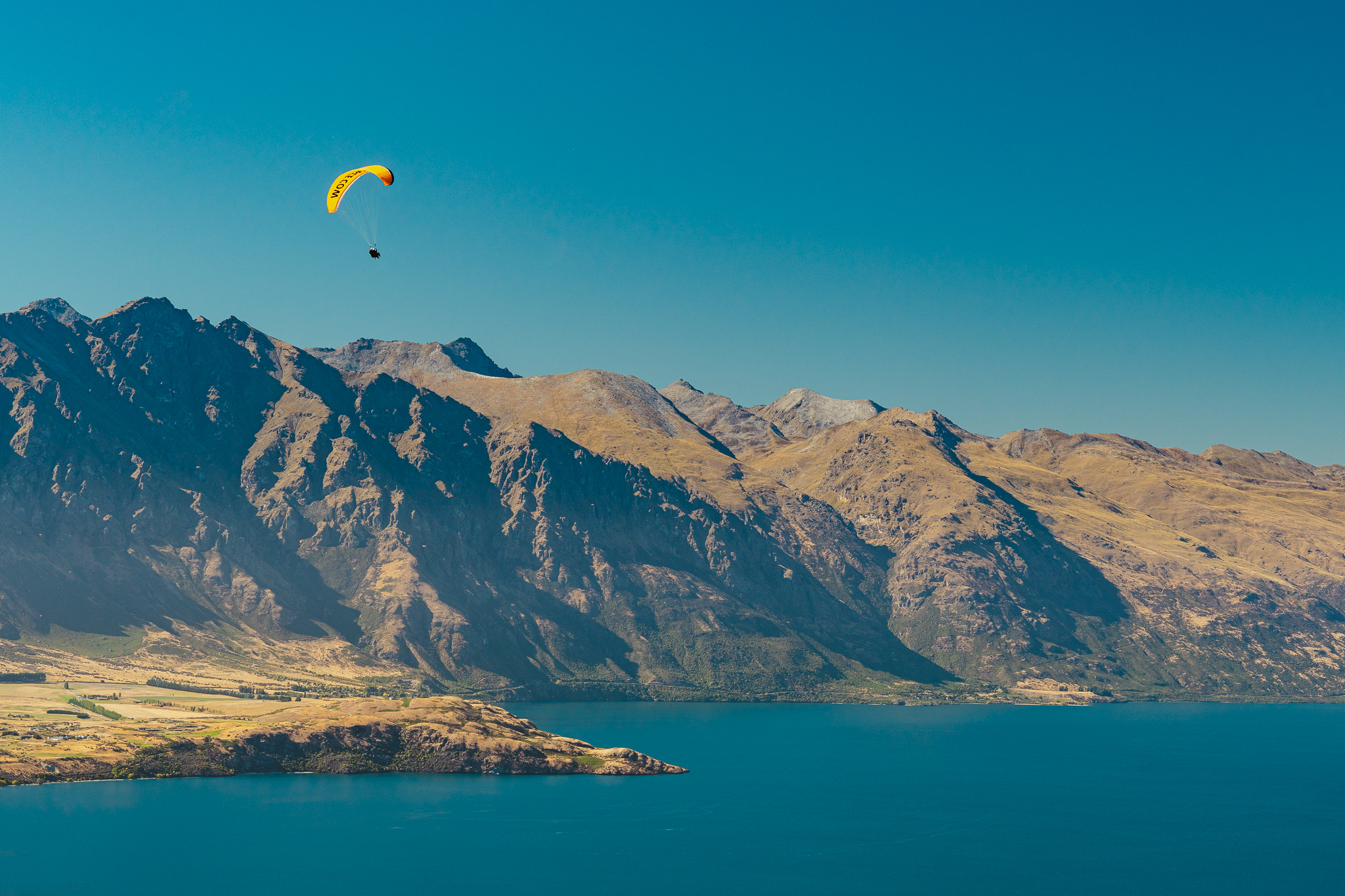 Aerial view of a lake with a mountainous backdrop, clear blue water, and a small town nestled along the shoreline under a bright blue sky.