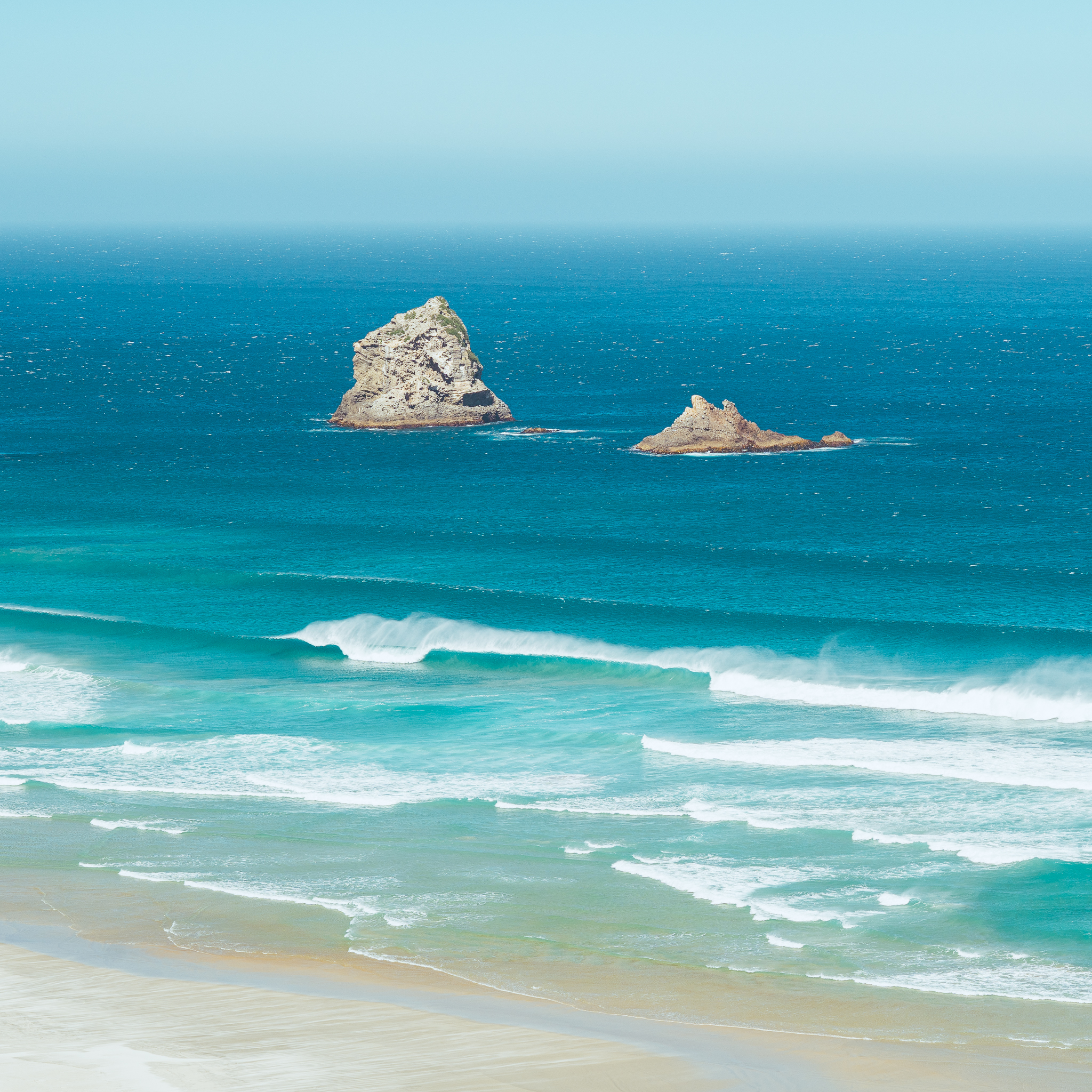 Two rocky islets in a vibrant blue ocean, with waves gently crashing onto a sandy beach under a clear sky.