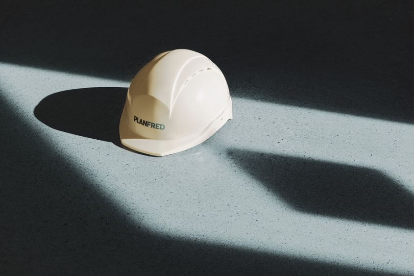 Weißer PLANFRED-Schutzhelm auf hellem Terrazzoboden, angestrahlt von einem Streifen Sonnenlicht mit langen Schatten