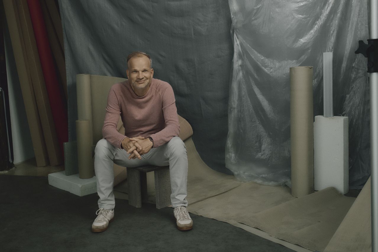 Maximilian Schmid im rosa Pullover sitzt lächelnd auf einem Hocker, umgeben von Papierrollen und Planen im Fotostudio