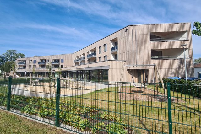 Holzfassade mit Spielplatz, Nestschaukel und Grünzaun bei blauem Himmel