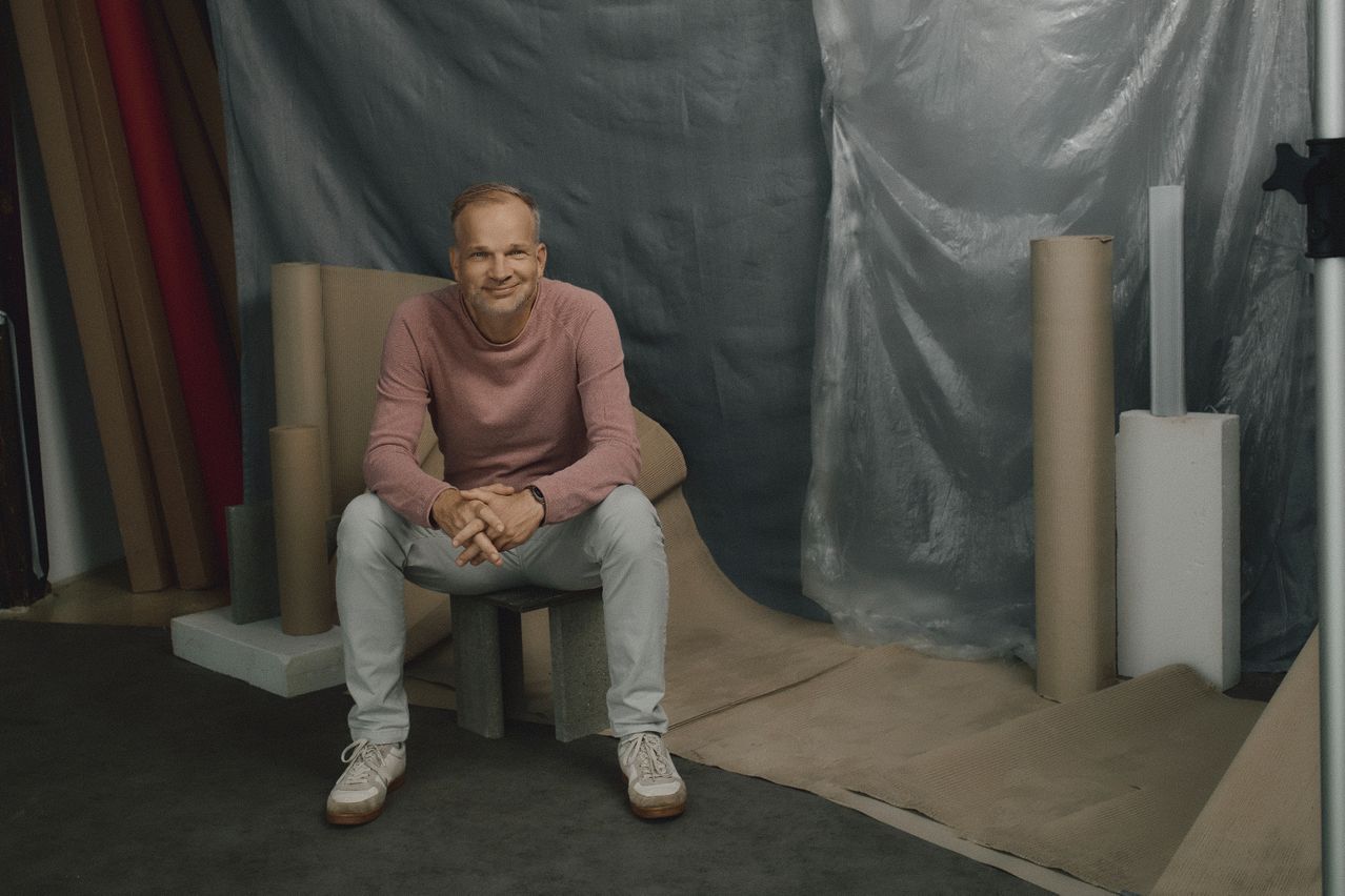 Maximilian Schmid im rosa Pullover sitzt lächelnd auf einem Hocker, umgeben von Papierrollen und Planen im Fotostudio