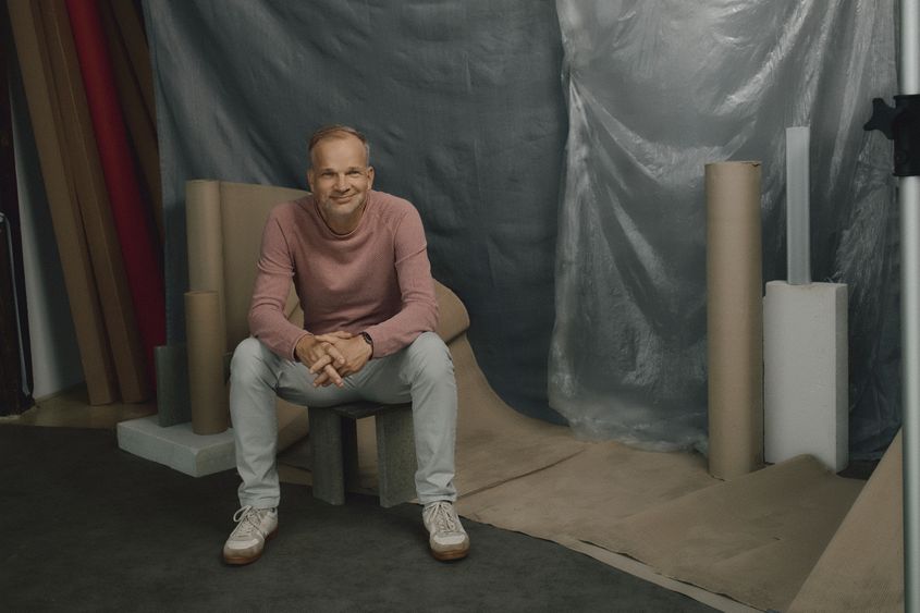 Maximilian Schmid im rosa Pullover sitzt lächelnd auf einem Hocker, umgeben von Papierrollen und Planen im Fotostudio