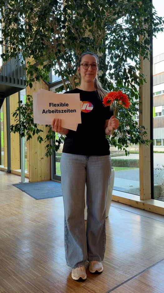 A person in a black shirt and jeans holds a sign reading "Flexible Arbeitszeiten" and flowers, standing in a modern, sunlit office space.