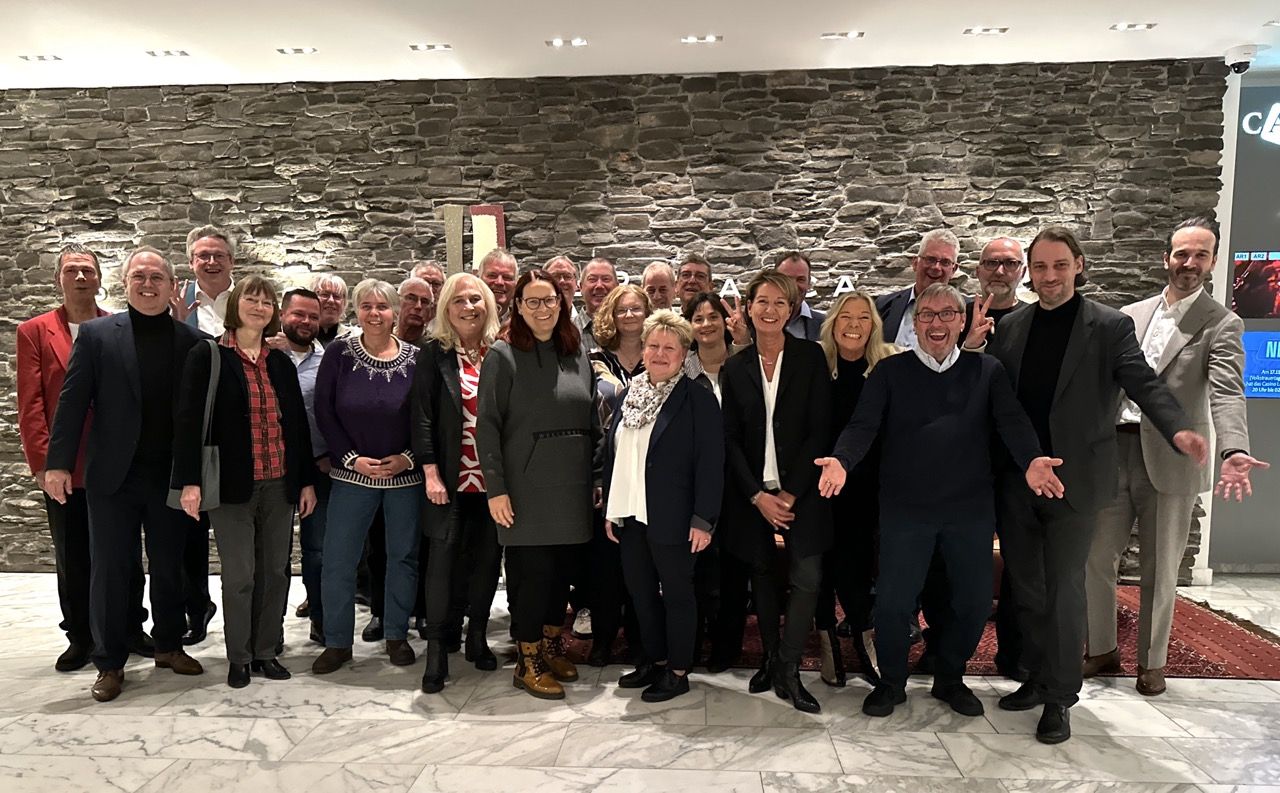 A group of people posing together indoors in front of a stone wall, some smiling and others gesturing with open arms.
