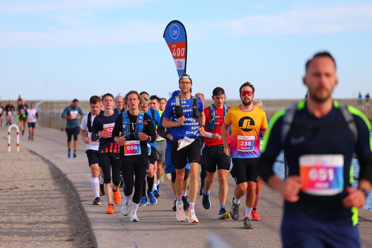 A group of runners participating in a marathon on a coastal path, led by a pacer holding a "5:00" sign.