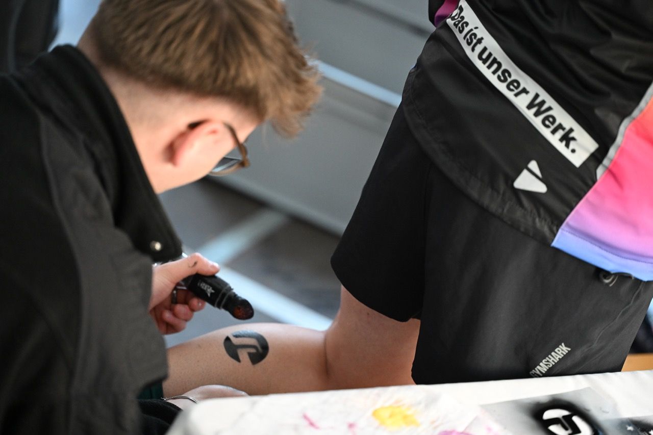 A person applies black ink to another's leg, creating a design. The person getting inked wears shorts with text and a logo visible.