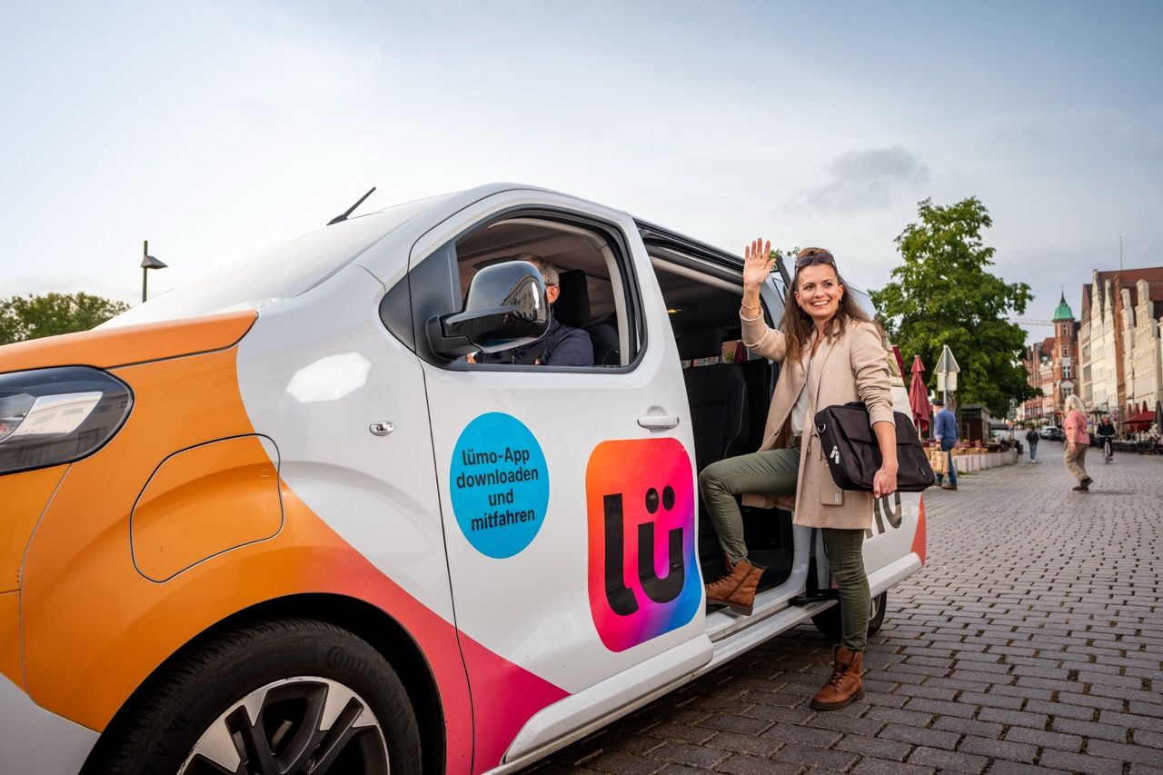 Woman waving and boarding a colorful Lümo van on a cobblestone street, with a driver inside and trees in the background.