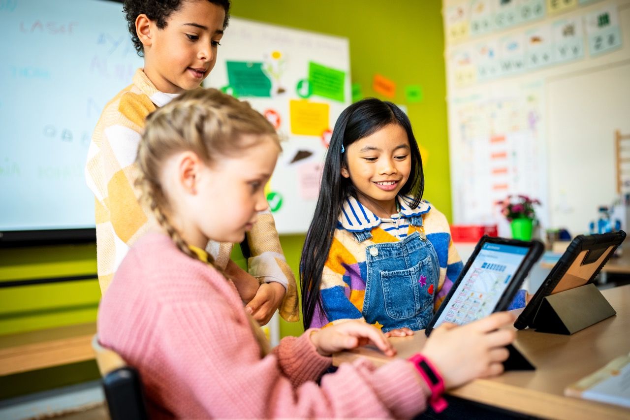Three children in a classroom use tablets, engaged and smiling. The background shows a whiteboard and colorful posters on green walls.