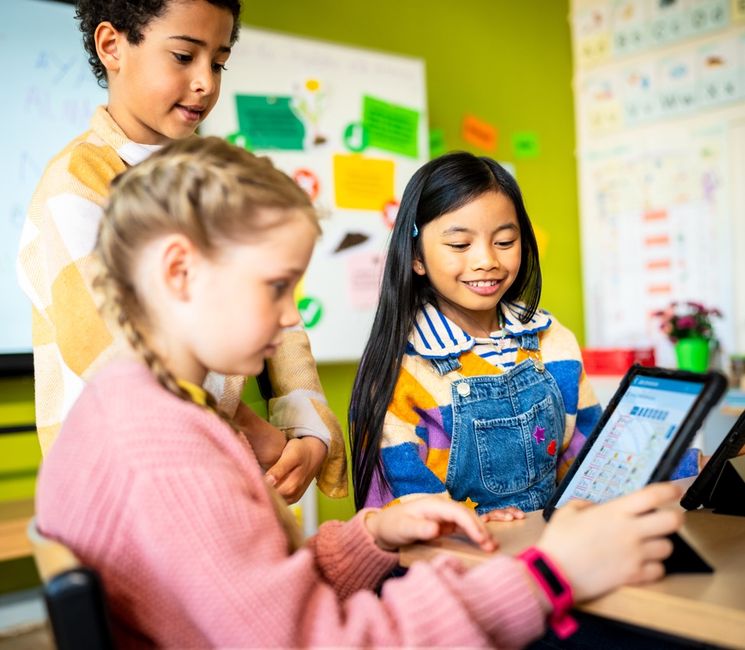 Three children in a classroom use tablets, engaged and smiling. The background shows a whiteboard and colorful posters on green walls.
