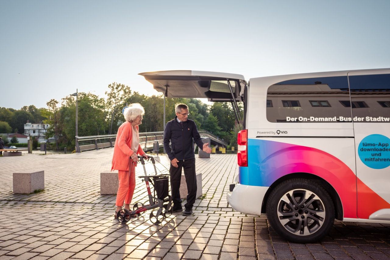 An elderly woman with a walker and a man stand near a colorful on-demand bus with an open rear door in a sunlit urban area.