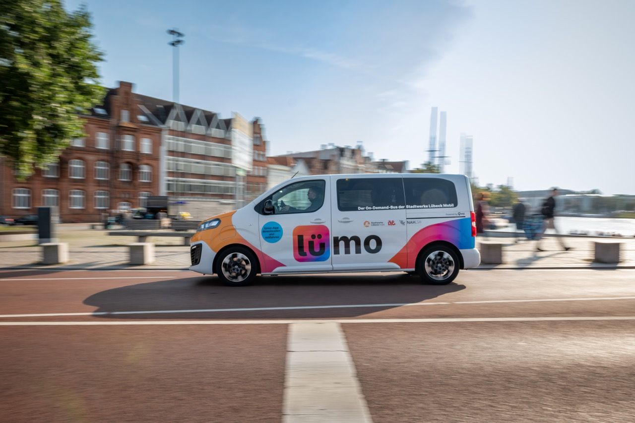Colorful van with "umo" logo driving on a city street, with brick buildings and trees in the background under a clear blue sky.