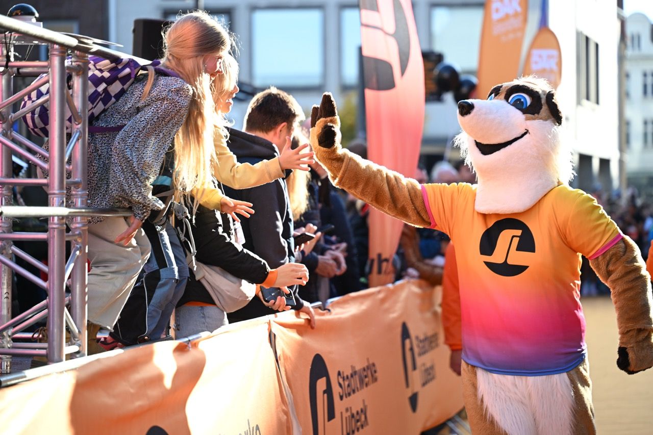 Mascot in a colorful shirt high-fives a child over a barrier, surrounded by a crowd and orange banners at an outdoor event.