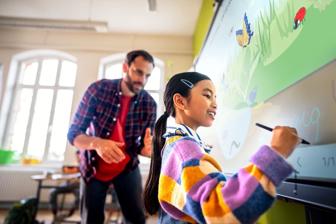 A young girl draws on an interactive whiteboard in a classroom, while a teacher in a plaid shirt observes and guides her.